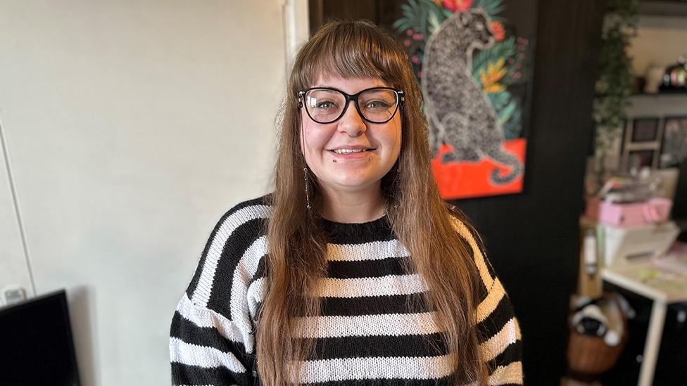 A young woman with large glasses and long brown hair with a straight fringe wears a black and white-striped jumper. She's standing in a living room with a tapestry hanging on the wall depicting a big cat of some sort. In the corner we can just make out sewing supplies.