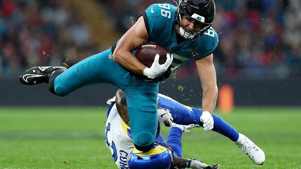 American football player in teal uniform runs with the ball while being tackled by an opponent in white and blue on a grassy field during a game.
