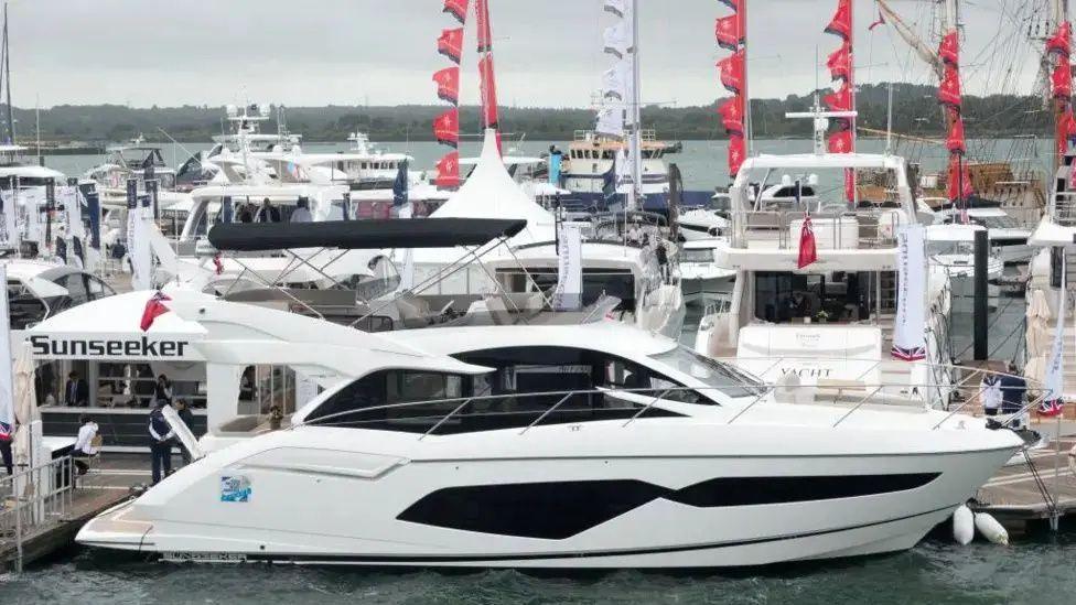 A luxury yacht is pictured docked in a busy harbour. On the jetty a gazebo says 'Sunseeker' on it