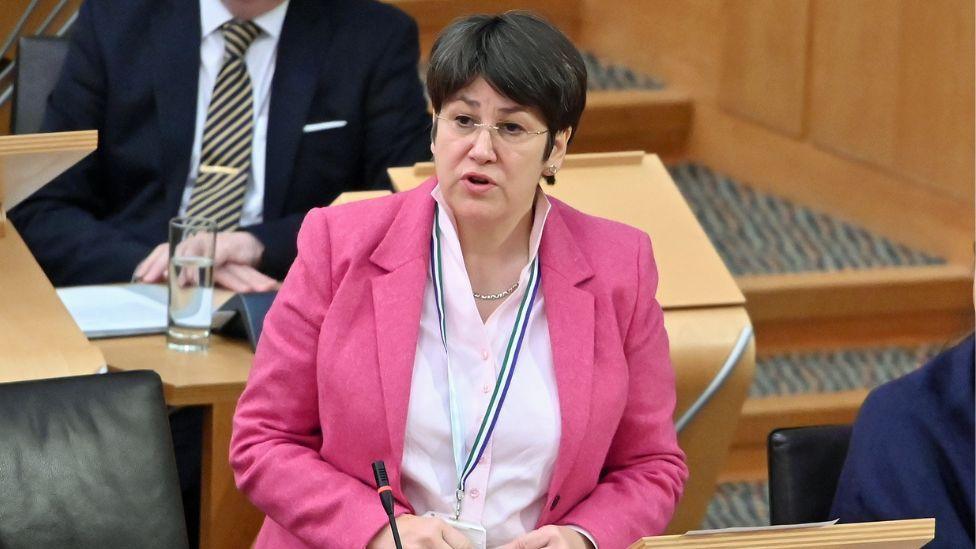 Tess White, a dark haired woman wearing a pink suit, speaks in the Scottish Parliament. She is visible from the waist up and is standing at a podium with a microphone in front of her. 