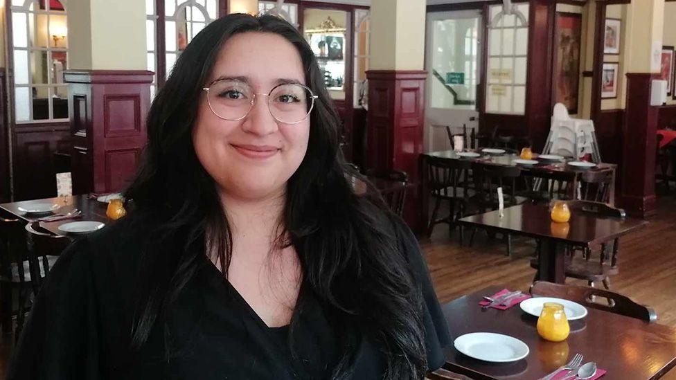 Natasha Khan, smiling, wearing round glasses, standing in a restaurant with a view of dining tables behind her