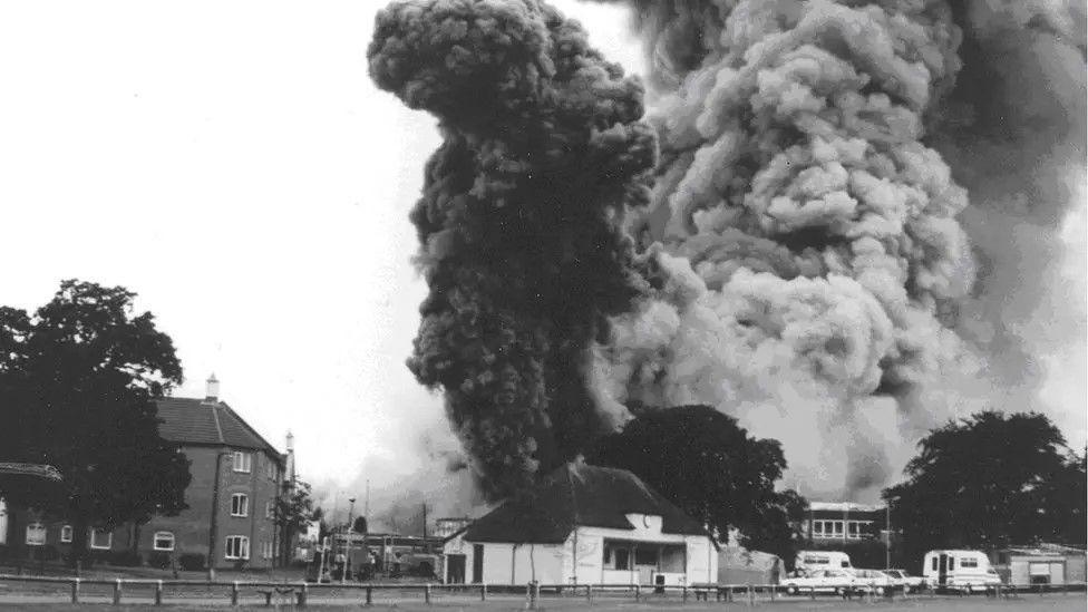 A black and white image of a huge fire; there are large plumes and clouds of thick black and grey smoke billowing from a barely visible building.
