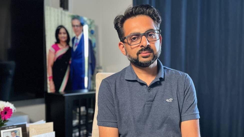 Miten Patel poses for a picture. He wears glasses and wears a blue polo shirt. A portrait of what appears to be his parents is seen in the background of the room.