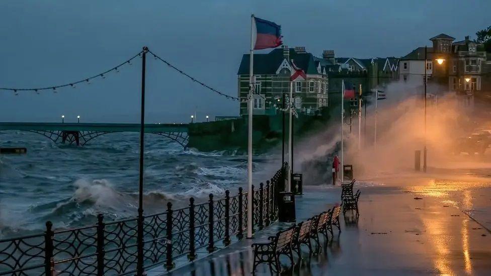 Large waves and a high tide batter the promenade on Clevedon sea front. The tide is very high up against the piece and flags on flag poles are shown blowing in the wind. Spray from the sea is seen over the pavement and road. 