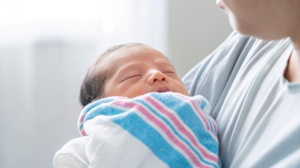 A newborn baby wrapped in a blue, white and pink blanket, held be a woman wearing a grey top.
