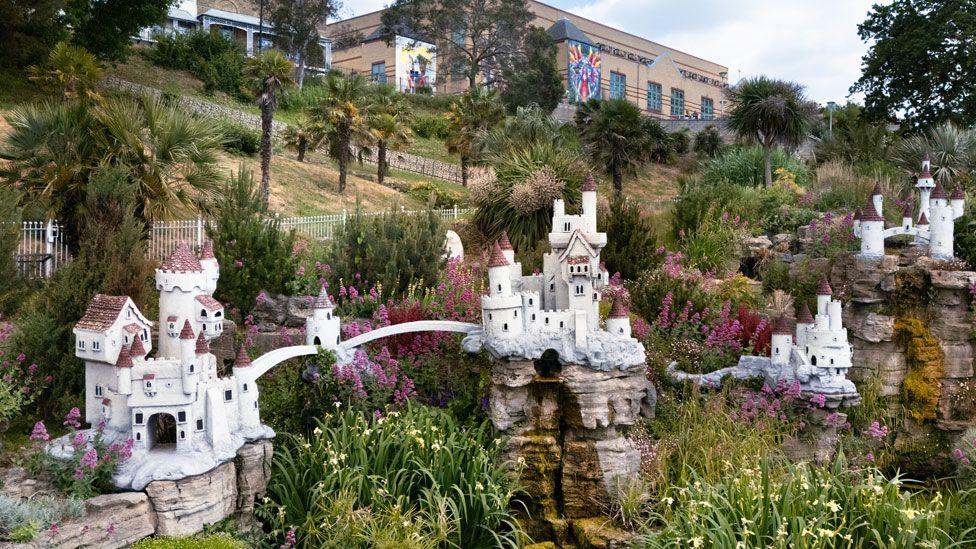 The Shrubbery, Royal Terrace, Southend-on-Sea, Essex. A general view of The Fairy Castle, Never Never Land, showing four linked white red-roofed turreted castles, linked by bridges. Pink and yellow flowered plants are growing in between. Beyond them is a slope with trees, rising up to buildings. 
