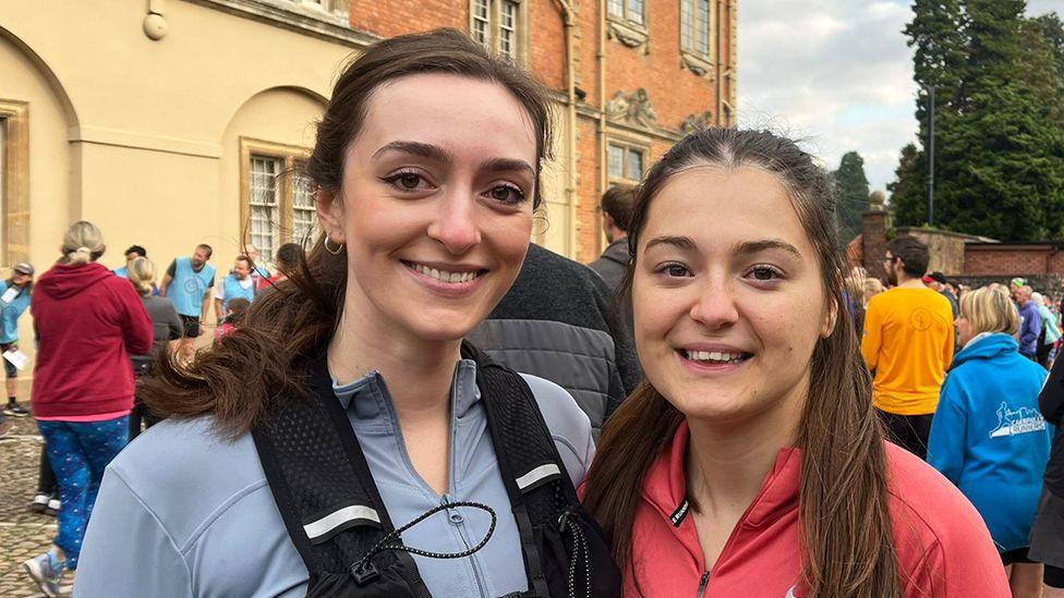 Two friends, Laura and Abbey, smiling next to each other. Abbey is wearing a running vest with blue fleece while Laura is wearing a red top. They are at a Parkrun event in Wales.