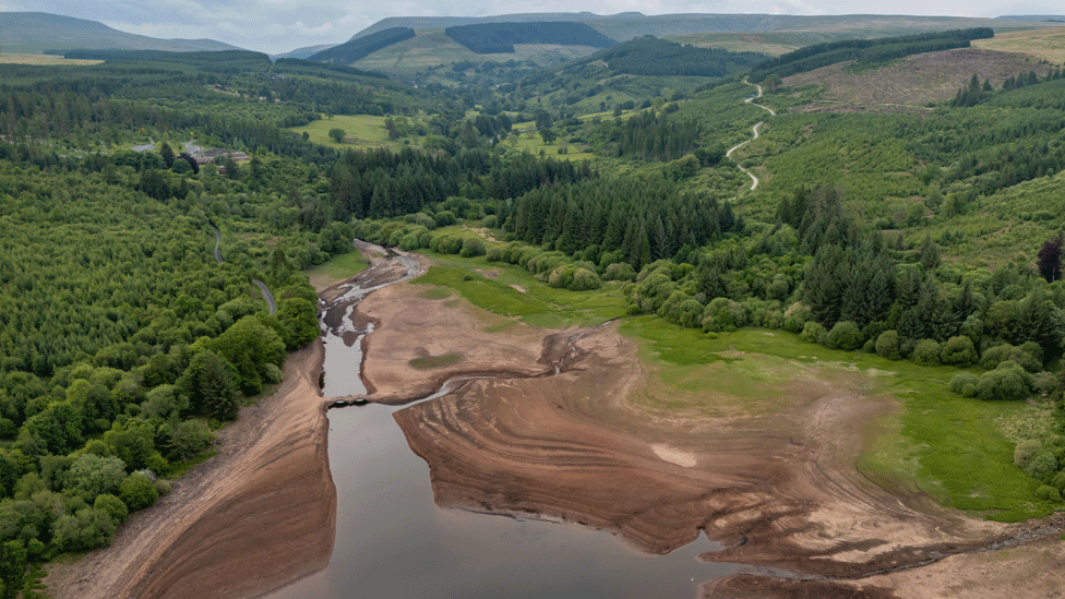 Aerial drone view looking down on the bridge and a wider dry expanse of the bed of the reservoir, surrounded by trees
