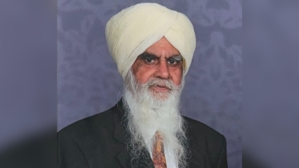 Bhai Joginder Singh wearing a dark suit, orange tie and a cream-coloured turban. He has a long white beard.