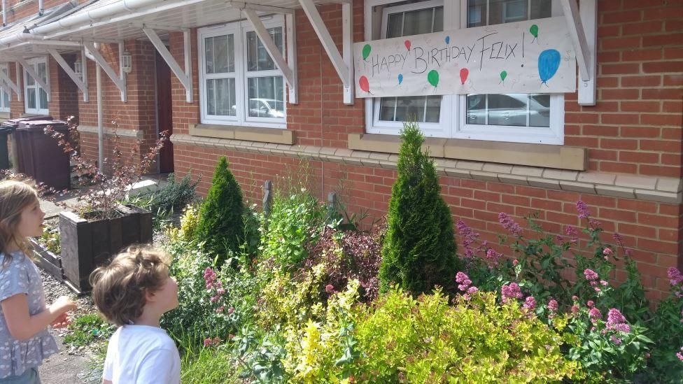 A young boy and girl stand in front of a front garden and look at a colourful sign that says "happy birthday Felix"