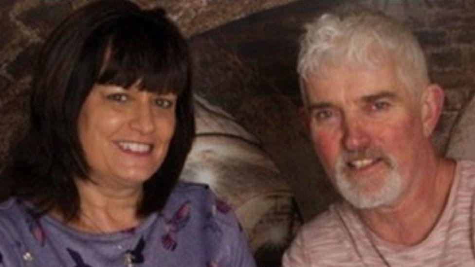 Maria and Nick Evans sitting next to each other, smiling at the camera. She has shoulder-length hair and is wearing a purple top. He has white hair and a beard and is wearing a pink T-shirt