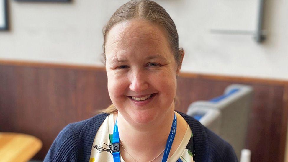 Head-and-shoulders image of a smiling Catherine Makin. She has long brown hair tied back in a ponytail with a centre parting. Smiling for the camera, she is wearing a cream top with a navy cardigan on top.