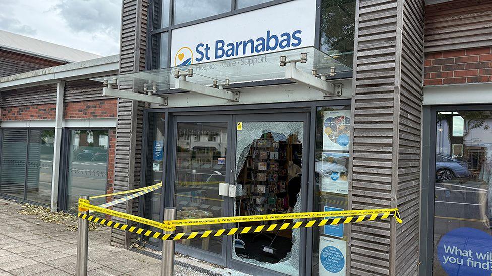 The outside of the St Barnabas shop showing the right-hand front door completely smashed there is yellow and black tape around the entrance