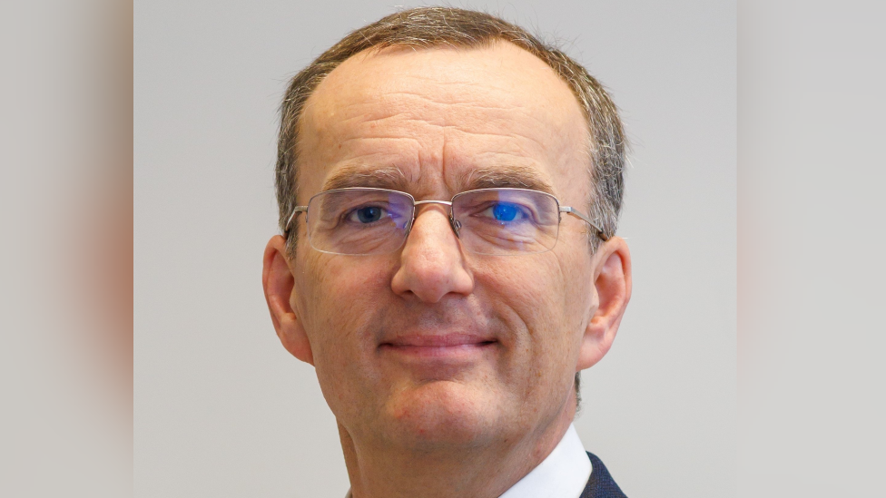 A man with short grey hair and glasses, wearing a collared white shirt and smiling at the camera.