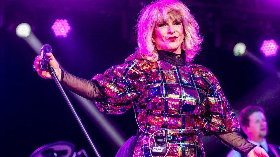 Toyah wearing a square-pattern sequinned dress holding a microphone to her side