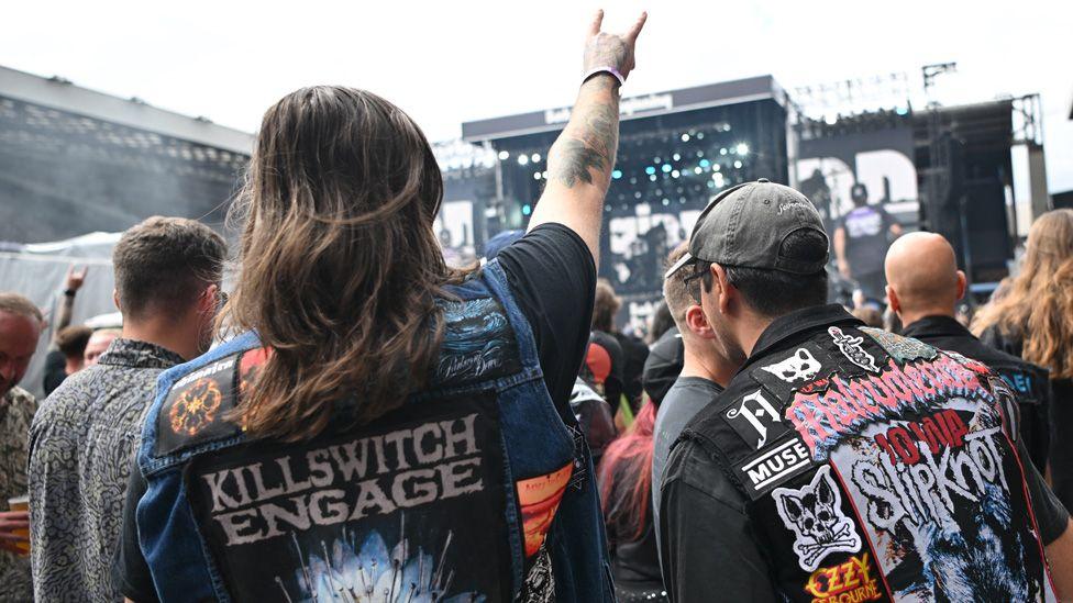 Two fans seen from behind on the packed pitch in the stadium, both wearing jackets with band logos on the back, and one with a hand in the air making the rock hand sign.