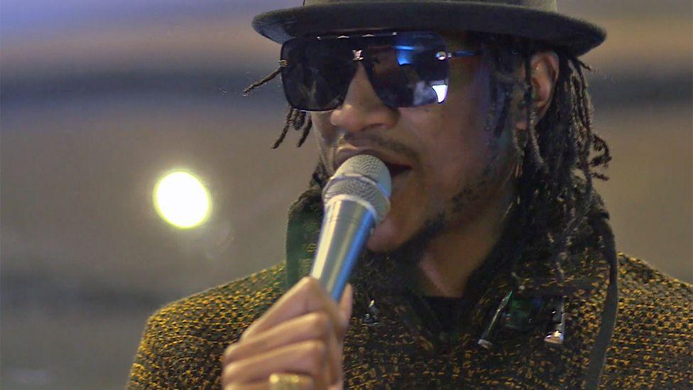 Jah Prayzah, wears shades, a black hat and in a yellow tweed-like top, as he sings holding a microphone in a BBC studio in 2021