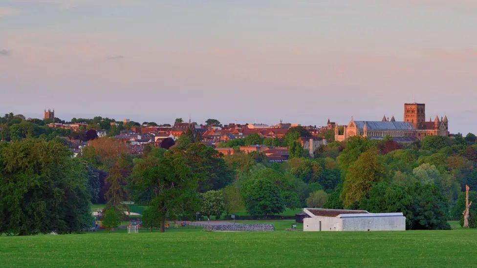 In the foreground a park with green field and trees, in the background a cathedral and red brick rooftops