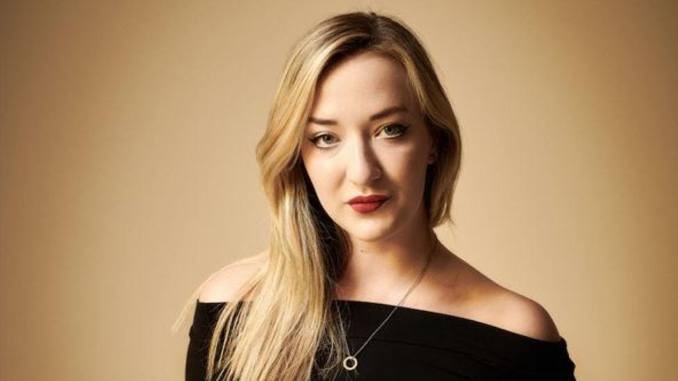 Woman with long blonde hair and dark top, wearing a necklace, looking at camera.