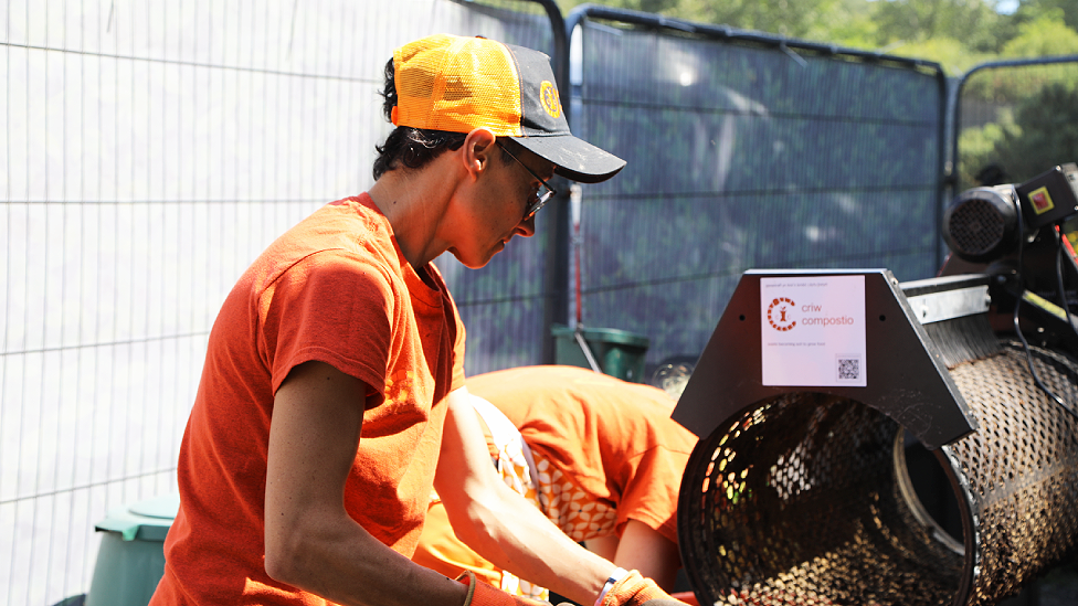 ffin with an orange and black baseball style cap on and an orange Criw Compostio t-shirt. She is wearing glasses and appears to be holding something in her hands out of the picture, possibly a shovel, and putting something into the circular opening of a composting or shredding machine to her side, which has a Criw Compostio label on the top of it. She is photographed from the side and looking down at what she is doing. Behind her to the right Steph, also wearing orange, is bent over and her head is out of sight behind the machine. There is movable fencing behind them.