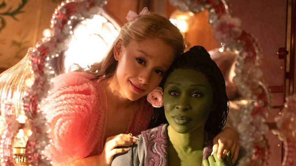 Ariana Grande and Cynthia Erivo stare into a pink mirror dressed as their Wicked characters Glinda and Elphaba.