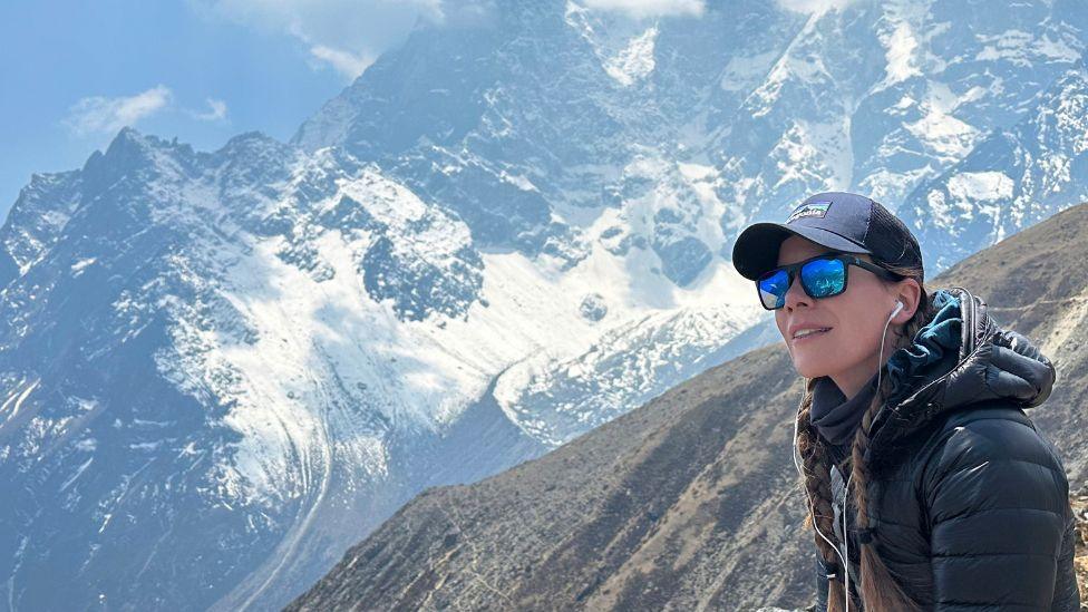 A woman wearing sunglasses and a baseball cap stands on a mountain side, with a snowy slope in the background