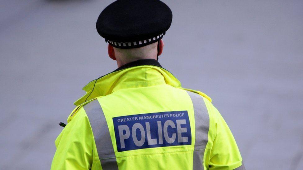 A police officer in a reflective yellow coat and wearing a black cap, with the words 'Greater Manchester Police' on the back, stands with his back to the camera