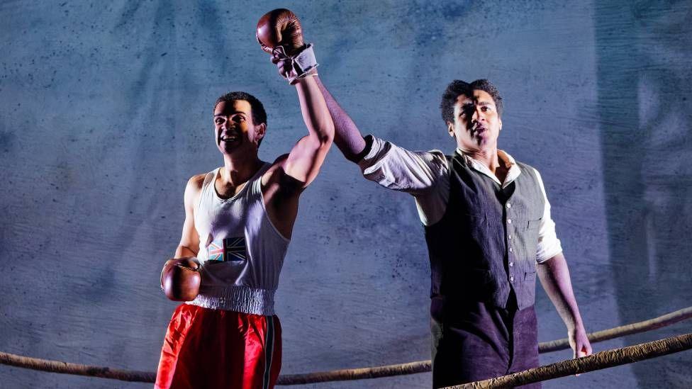 In a picture from the play, a boxer wearing a white vest, red shorts and red gloves has his arm held aloft, apparently in victory, by a man wearing a blue waistcoat and white shirt