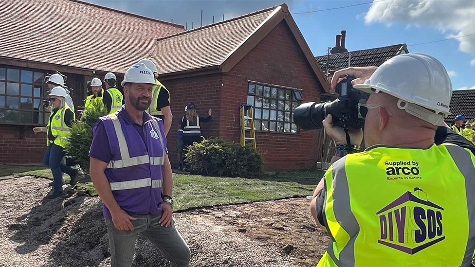 Nick Knowles in a purple high vis vest, purple polo shirt, grey jeans and white hard hat being photographed in front of a red brick bungalow by a man in a yellow high vis vest and white hard hat. In the background five workers walk across the front garden - a patch of grass in the middle of a trampled area of soil