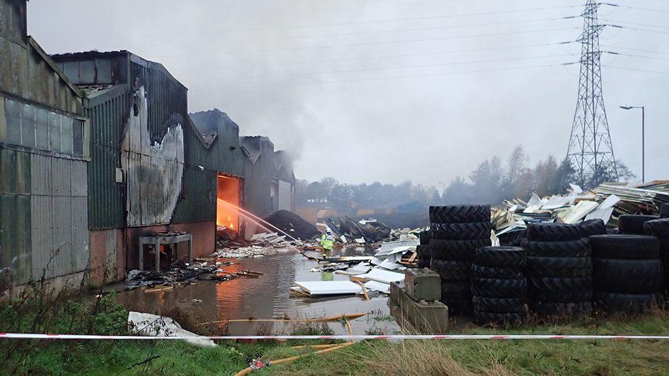 Wide view of the NWR site with the fire burning inside one unit, with water being poured onto the fire, tyres piled up to one side and what looks like an image of a giant chicken painted on the wall.