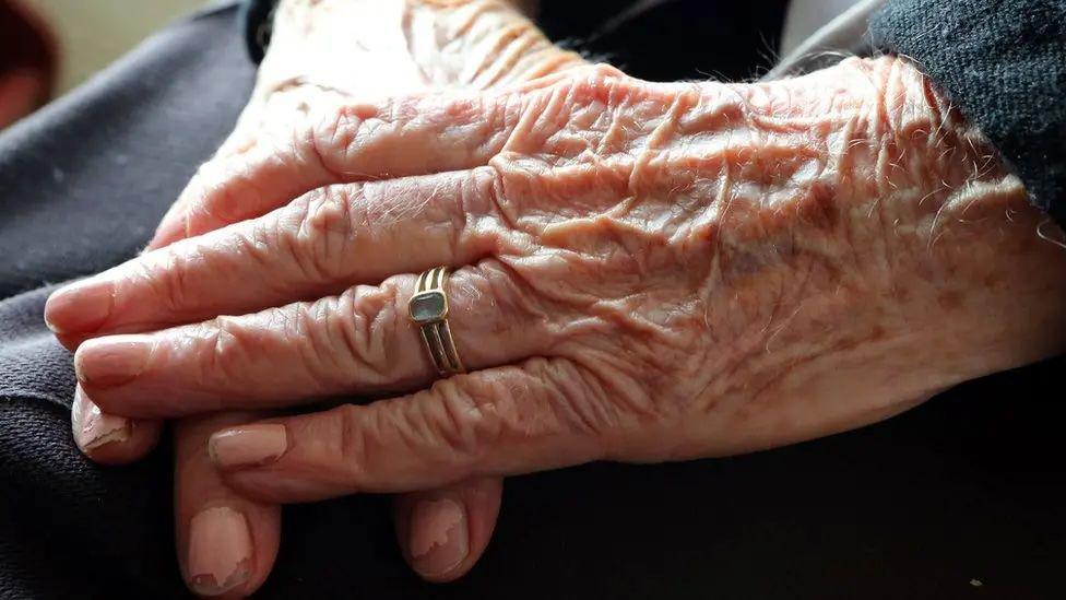 A closeup of the hands of an older person, with one over the other placed on their lap. There is a gold ring with a stone in it on the ring finger on the left hand, which is visible, and chipped pink nail varnish on the nails on both hands.