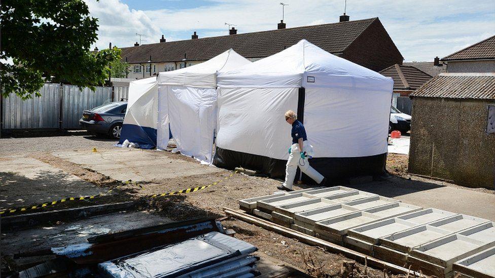 White tents in a residential car parking area being searched by forensic officers. The car park has a gravel surface with yellow and black tape across part of it. It lies in front of a row of houses