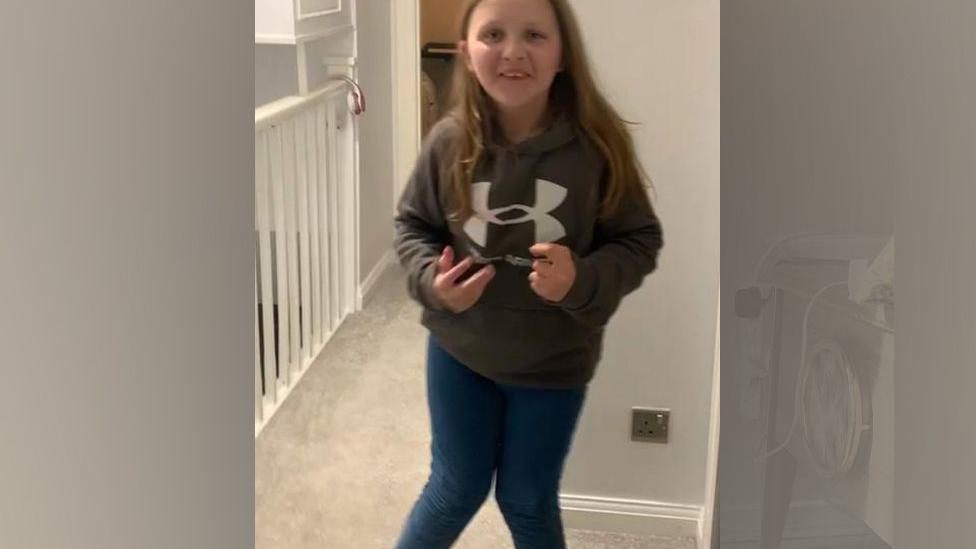 A young girl standing inside her house in a sweatshirt and jeans, showing that both her legs bend inwards at the knees. She is smiling at the camera.