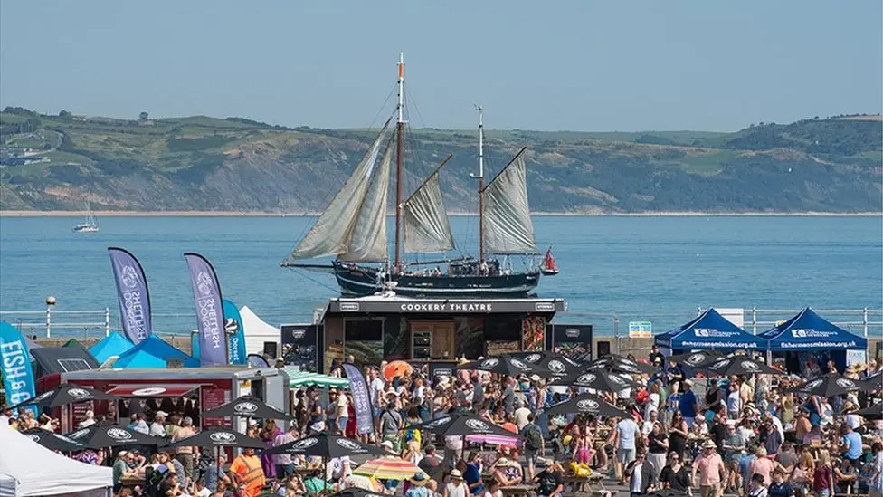 The festival is set up at Weymouth Peninsula. Crowds can be seen gathered around stalls and there is a ship with white sails in the sea in the background, with green hillside seen in the distance.