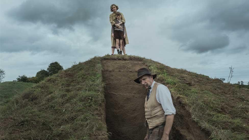 A still from the movie The Dig. Edith Pretty (Carey Mulligan) stands on top of a mounds with her arms around her son who is standing in front of her. Basil Brown (Ralph Fiennes) is at the bottom of the mound with his pipe in his mouth. A vertical section of grass has been removed from the mound - probably about a metre across and a couple few inches deep.