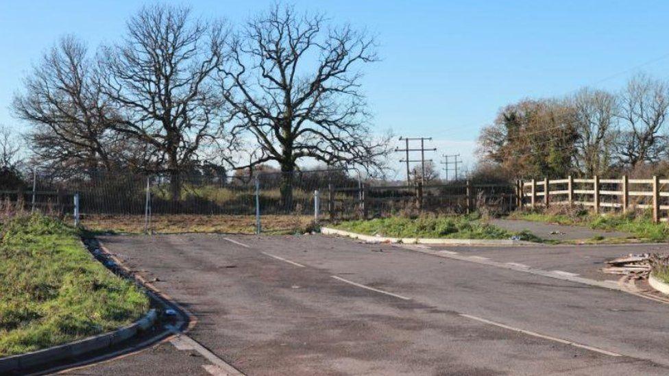 Road that stops at metal fencing with trees and fields beyond.