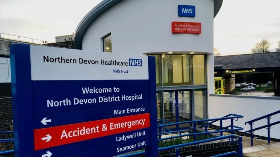 A blue, red and white  sign outside a hospital says: Northern Devon Healthcare NHS. Welcome to North Devon District Hospital. Arrows point toward the Main Entrance, Accident and Emergency and the Ladywell and Seamoor Units. Blur railings down steps go towards a glass sliding door.