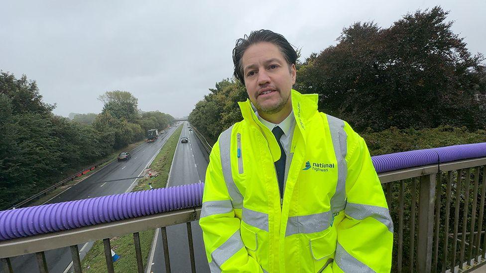 Graeme Woods stood on a bridge overlooking the A63 with dark hair and wearing a National Highways yellow high visibility jacket