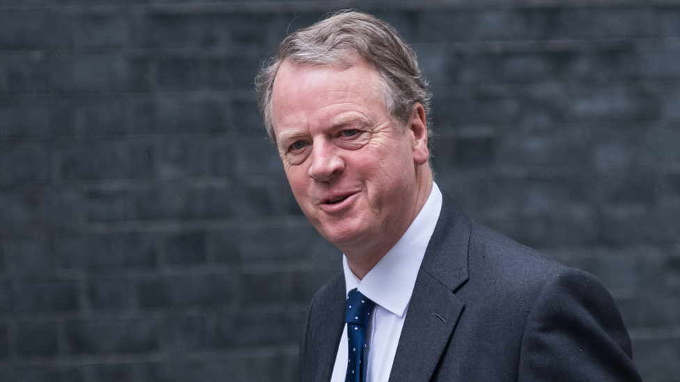 Alister Jack, who has dark grey hair, looks at the camera while walking in front of a grey brick wall. He is wearing a dark grey suit, white shirt and blue tie. 