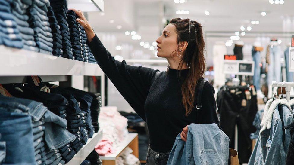 Young woman with brown hair in clothing store, looking at shelves stacked with jeans.