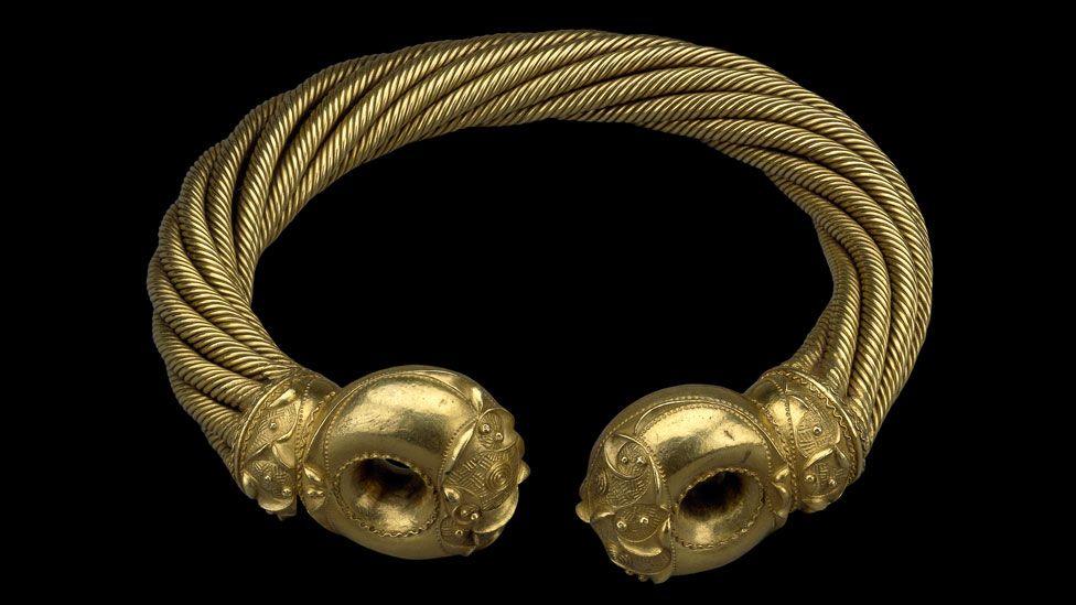 The Great Torc of Snettisham lying against a black background. It consists of ropes of twisted gold in a circle, ending in two highly decorated terminals facing each other with a gap between.