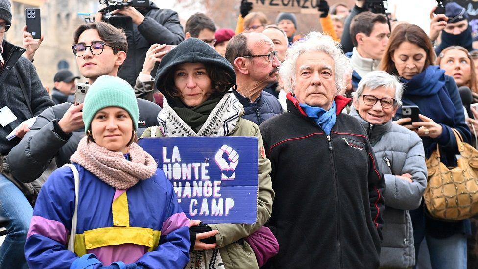 A group of people stand facing the camera. One at the front is colourfully dressed, and next to her a woman holds a sign in French that reads: Shame changes sides