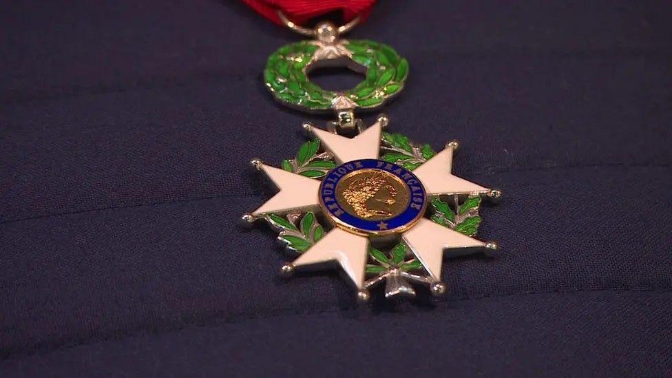 Close up of the Legion D'Honneur medal - a circular central blue ring medal with five white emanating two-pointed prongs, suspended from a green and silver laurel wreath and a red, watered silk ribbon