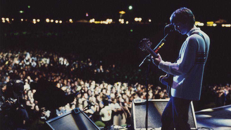 Noel Gallagher on stage at Knebworth with the crowd in the background and lit only by stage lighting