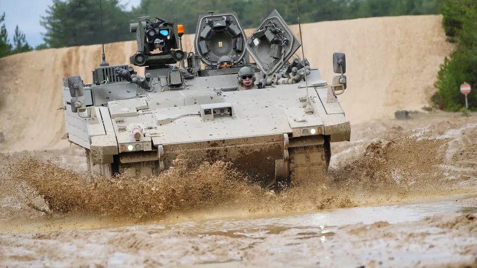An Ajax armoured vehicle being driven through a muddy terrain. Mud is being kicked up off the surface in all directions around the vehicle. Soldiers can be seen controlling the vehicle.