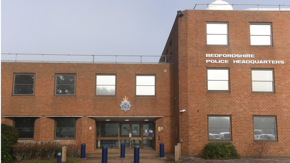 Front of Bedfordshire Police headquarters. A red-brick building with large windows and "Bedfordshire Police Headquarters" written on the front of it. It has a number of car bollards outside its front door. 