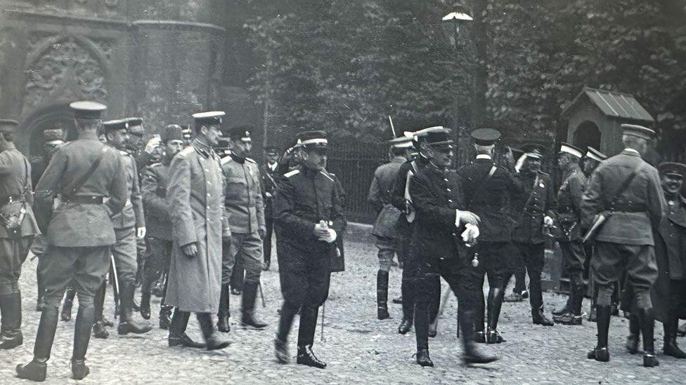 A black and white photo of at least 20 Army officers in a variety of uniforms milling around in front of a college entrance in Cambridge.
