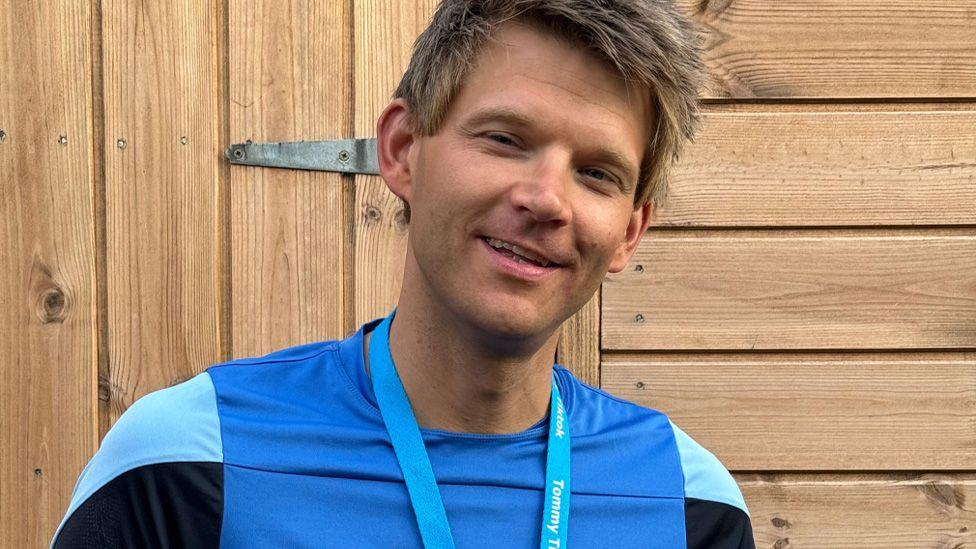 Tommy T in a blue sports top looking to camera. He has mousy brown spiky hair and is smiling. 