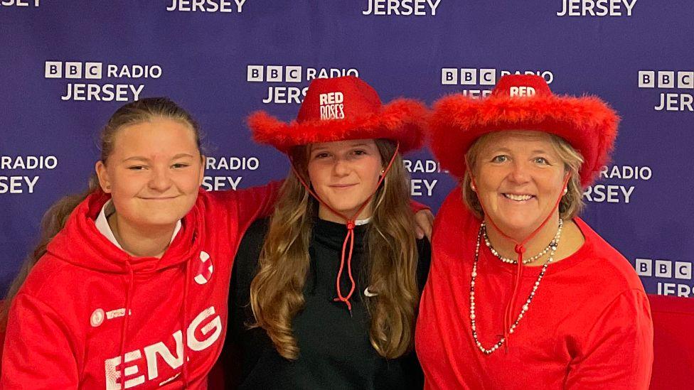 Alice, Maisie and Julia are sitting on a red couch in front of a backdrop with the BBC Radio Jersey logo repeated multiple times. The person on the left is wearing a red hoodie with 'ENG' written on it, the person in the middle is wearing a black hoodie and a red cowboy hat that says 'RED NOSE,' and the person on the right is wearing a red shirt and a large red cowboy hat. All three are smiling at the camera.