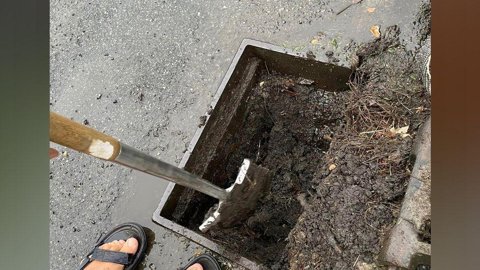 A spade digging inside a road drain which is full of mud and debris.